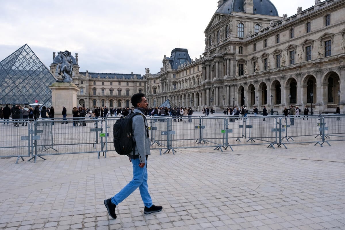 A man wearing a backpack walks in front of metal barriers in a large square backed by a glass pyramid and an ornate building. historic 