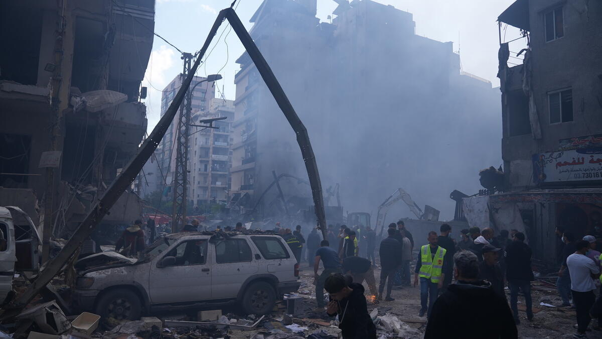 Crowds of people stand on a city street amid damaged buildings and vehicles seen through a haze of smoke