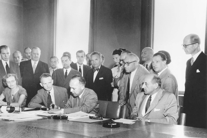 Back and white photo: A man signs the 1951 Refugee Convention, surrounded by a group of people.