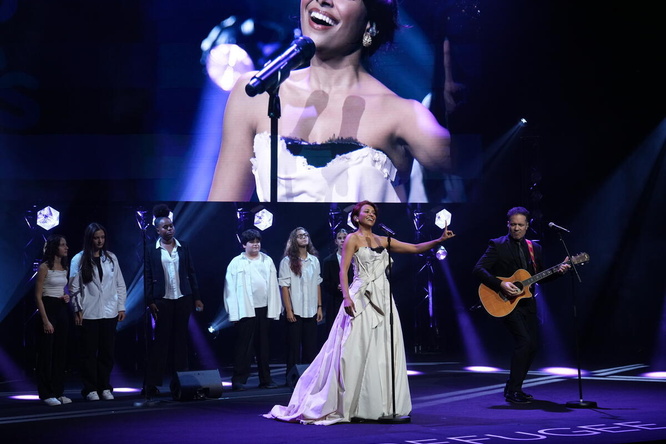 A woman performs onstage with a guitarist and backing singers in front of a giant screen