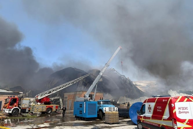 Black smoke surrounds a damaged warehouse, with emergency vehicles attending