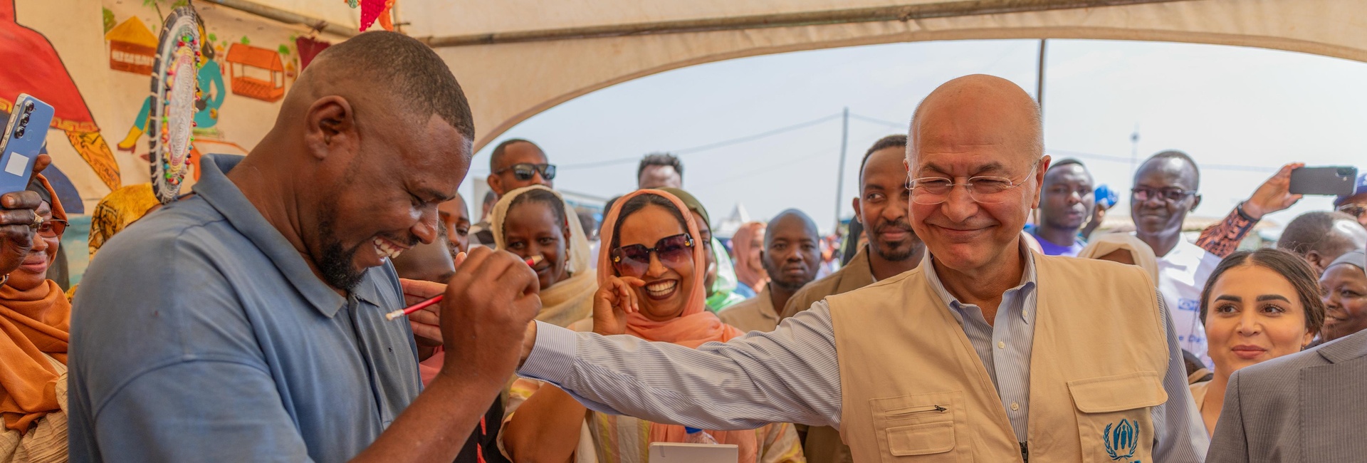 A smiling man drawing a pencil portrait on paper receives a pat on the shoulder from his smiling subject in a tent full of onlookers