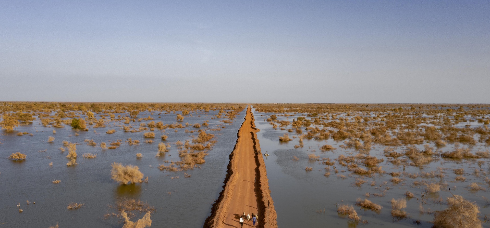 People walk along a diked road stretching to the horizon cutting through flood water
