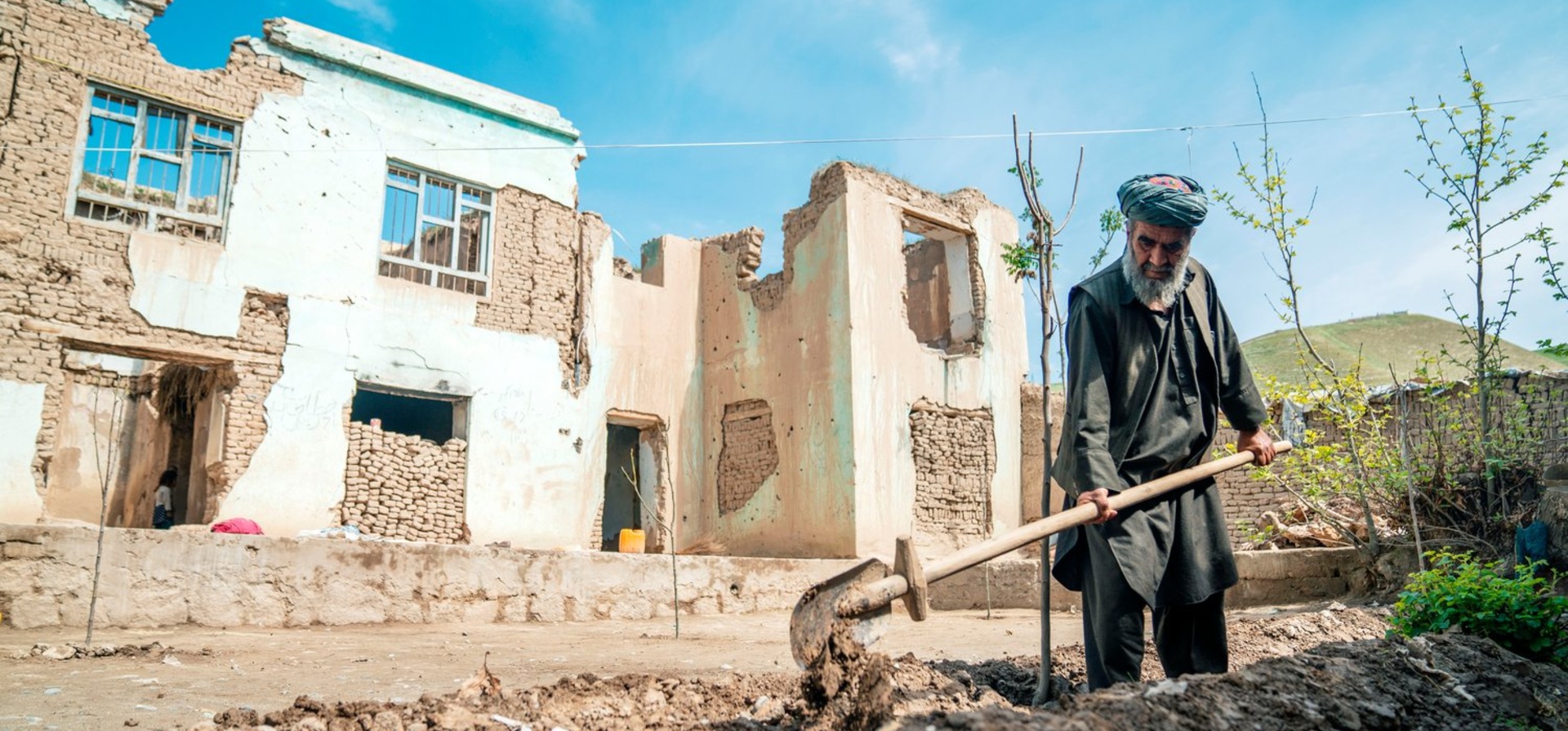 A man digs a trench for a tree in front of the ruins of a bombed-out house.