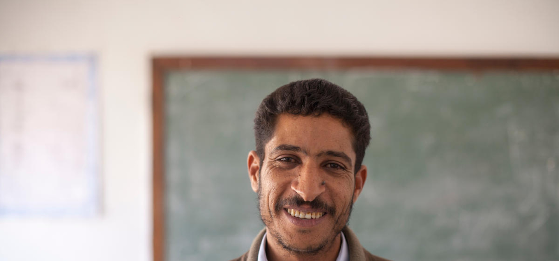 Portrait of a smiling male teacher, Mohammed, standing in front of a chalkboard in a classroom in Gwalish, Libya.