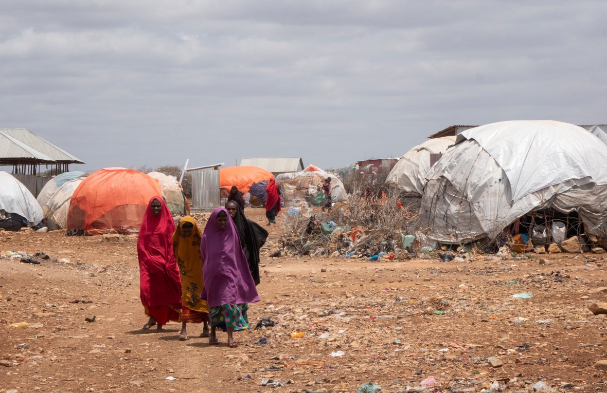 Four women walk through a camp made up of makeshift shelters.