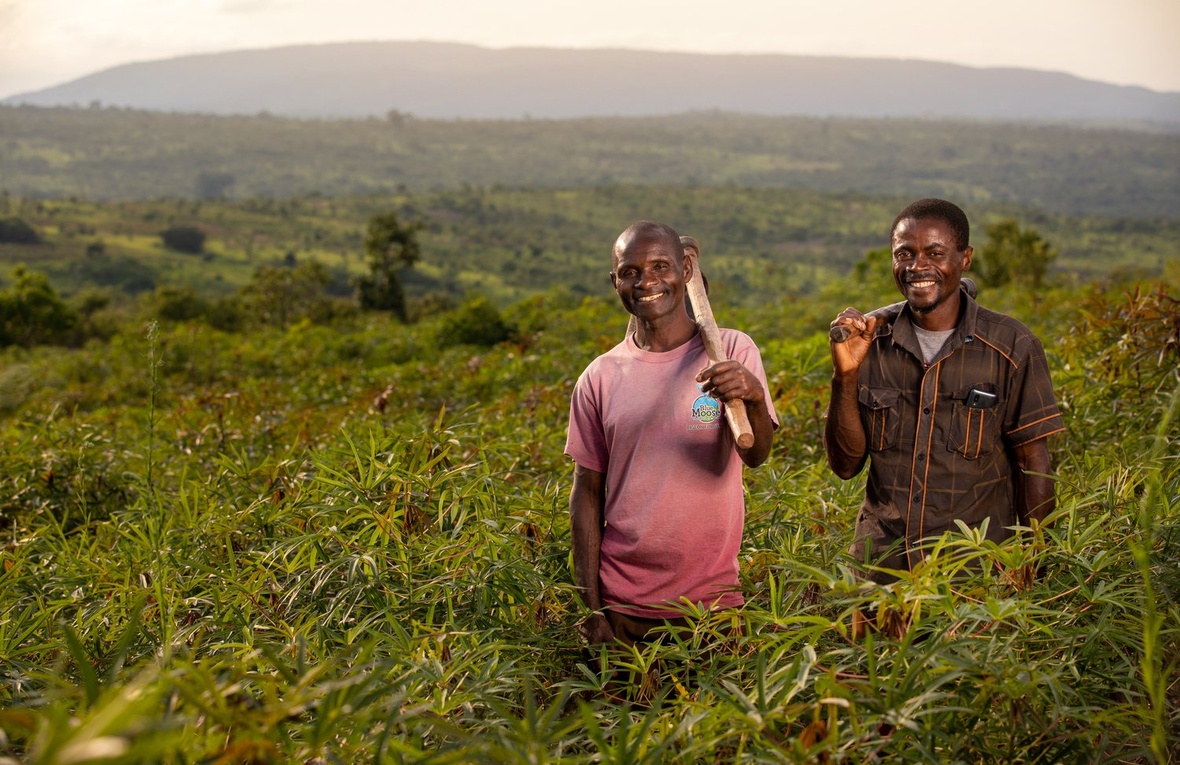 Two smiling men stand in a field holding farming tools