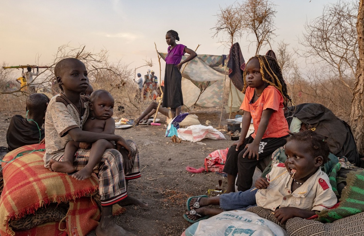 South Sudanese children sit in the bush with their mother standing in the background