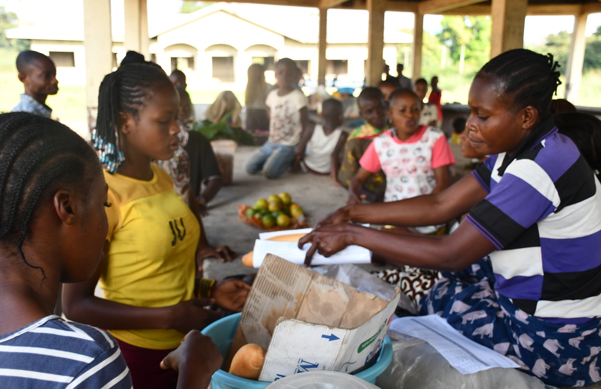 At a market, a woman hands over a small baguette wrapped in paper to a customer.