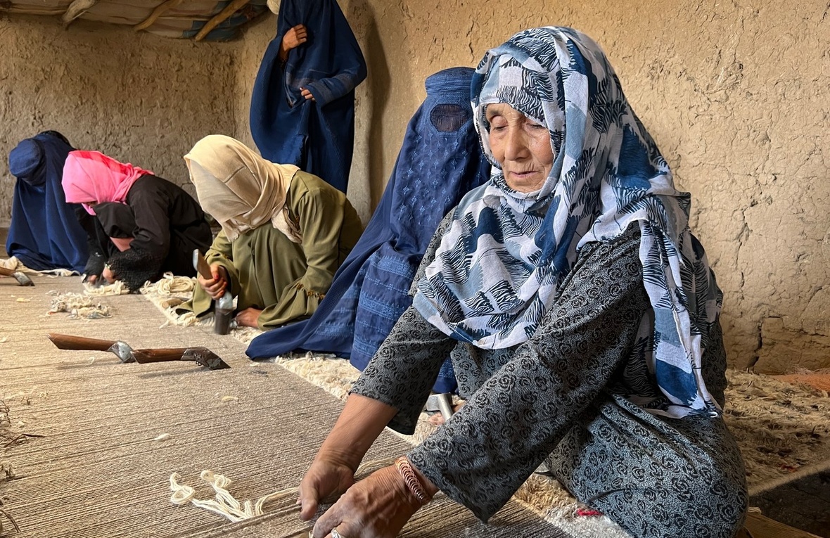 Several women sit on the floor of a hut weaving a large carpet.