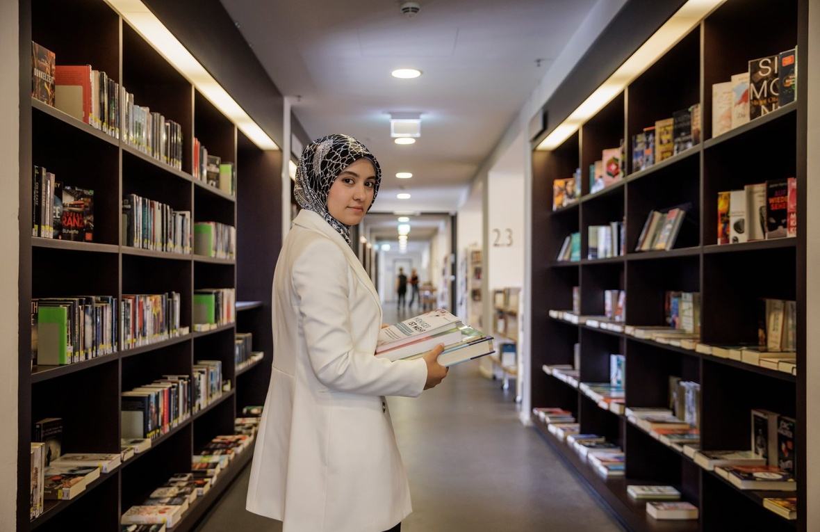 A woman in a lab coat stands between stacks of books in a library.
