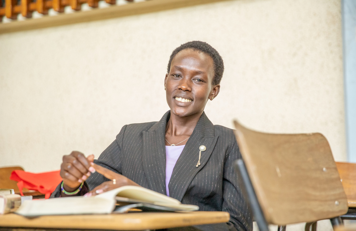 A woman in a suit jacket sits behind a desk in a lecture room.