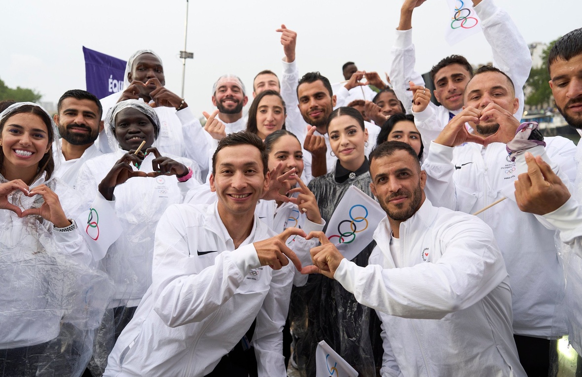 A group of young men and women wearing white tracksuits make heart emojis with their fingers.