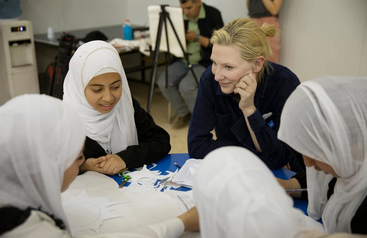 A smiling woman and four girls sit around a table holding scissors and paper 