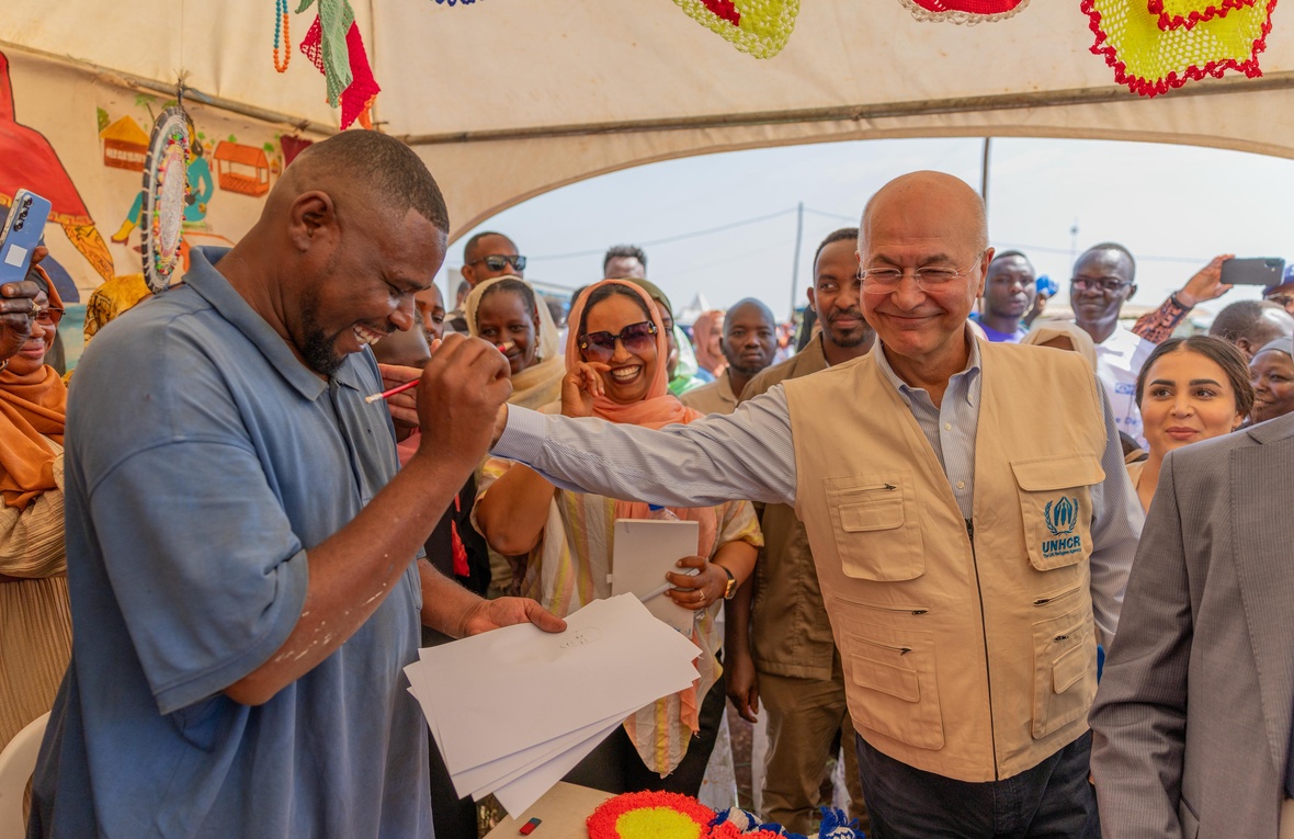 A smiling man drawing a pencil portrait on paper receives a pat on the shoulder from his smiling subject in a tent full of onlookers