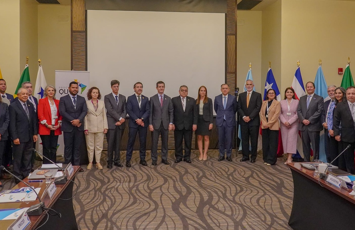 Un grupo de 18 hombres y 11 mujeres con vestimenta formal, de pie en una sala de juntas, detrás de ellos se encuentran 13 banderas coloridas de diferentes países