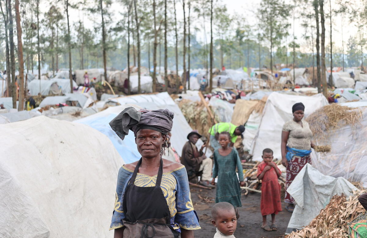 A woman stands in front of several shelters made of sticks and tarpaulins