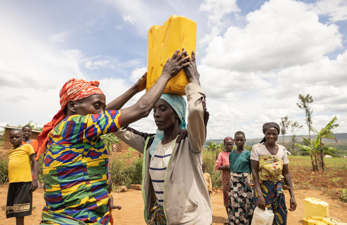 Women in colourful clothing, in a dry landscape, collect water in cannisters, carrying them on their heads or in their hands. 