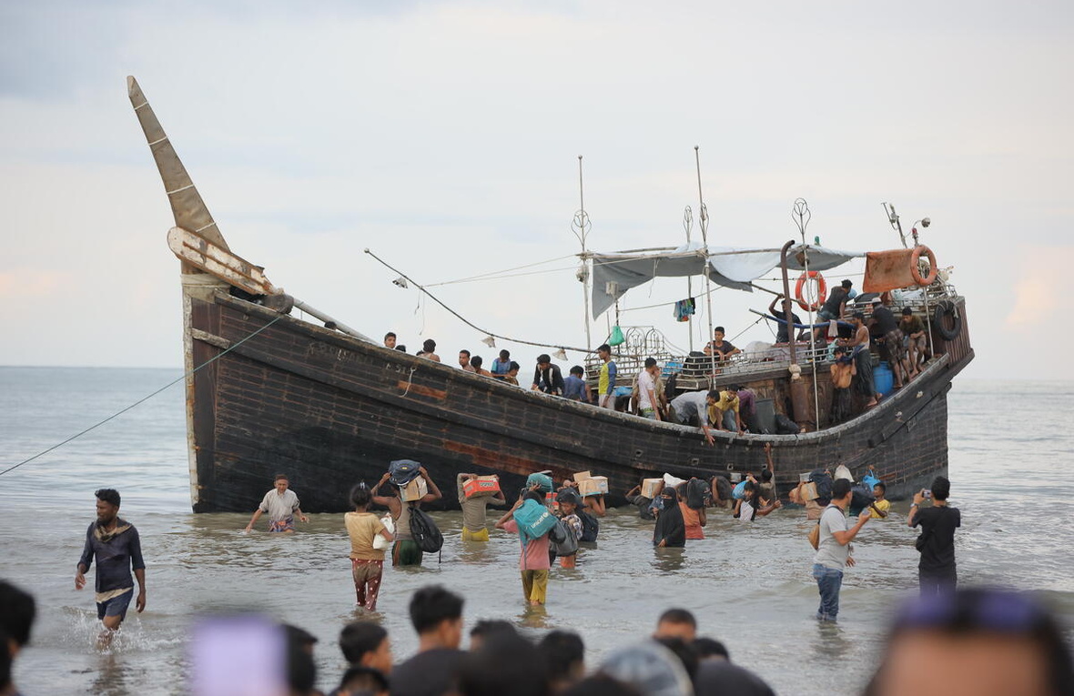 A group of people carrying belongings stand in the shallows alongside a wooden boat carrying refugees.