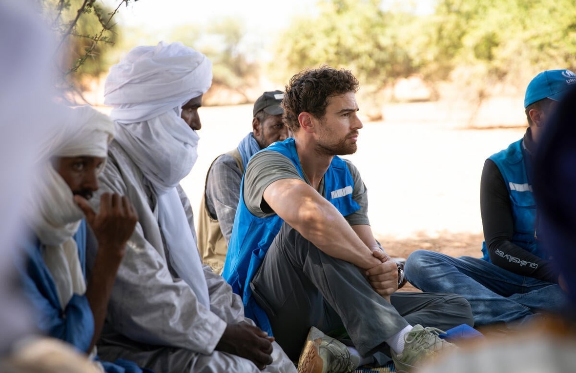 A man wearing a blue UNHCR vest sits on the ground outside next to men wearing white headscarves.