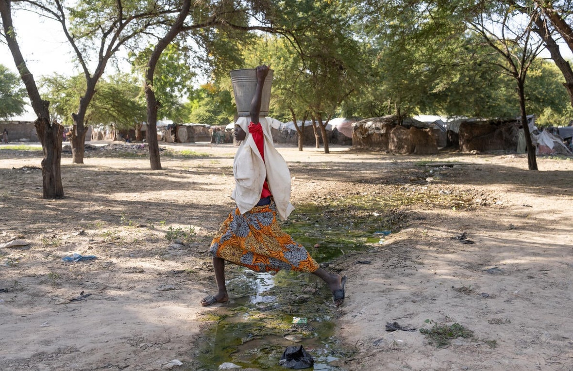 A girl holding a bucket on her head steps over a stream running through a camp.