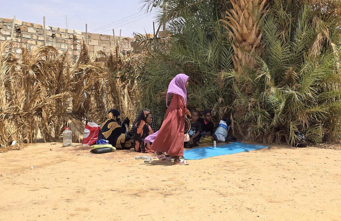 A Sudanese woman walks by a group of people sitting in the shade of some large trees in a dry environment.