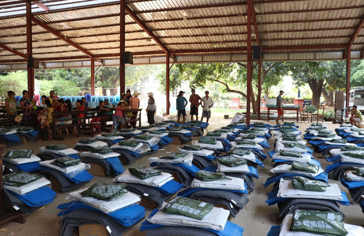 Rows of emergency supplies are laid out on the ground in a large shelter, where small groups of people gather at the back.