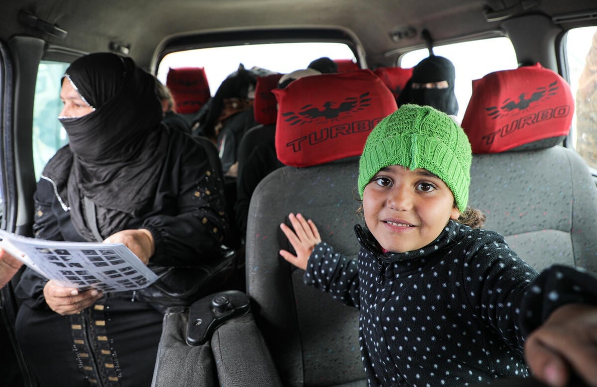 A young girl wearing a knitted green hat sits inside a minibus with women passengers sitting in the seats behind her.