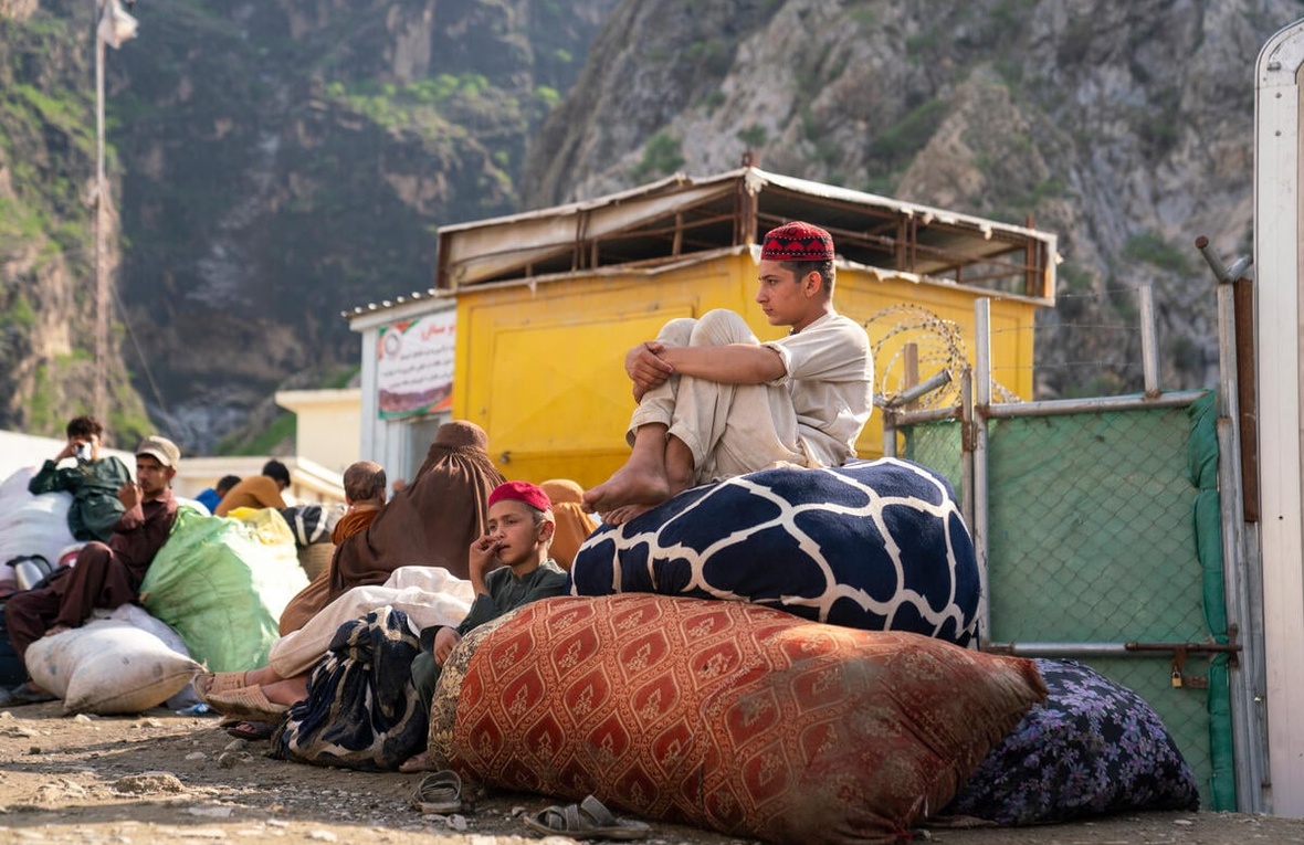 Two brothers wait at a bus stop in front of the mountains, sitting on large piles of belongings.