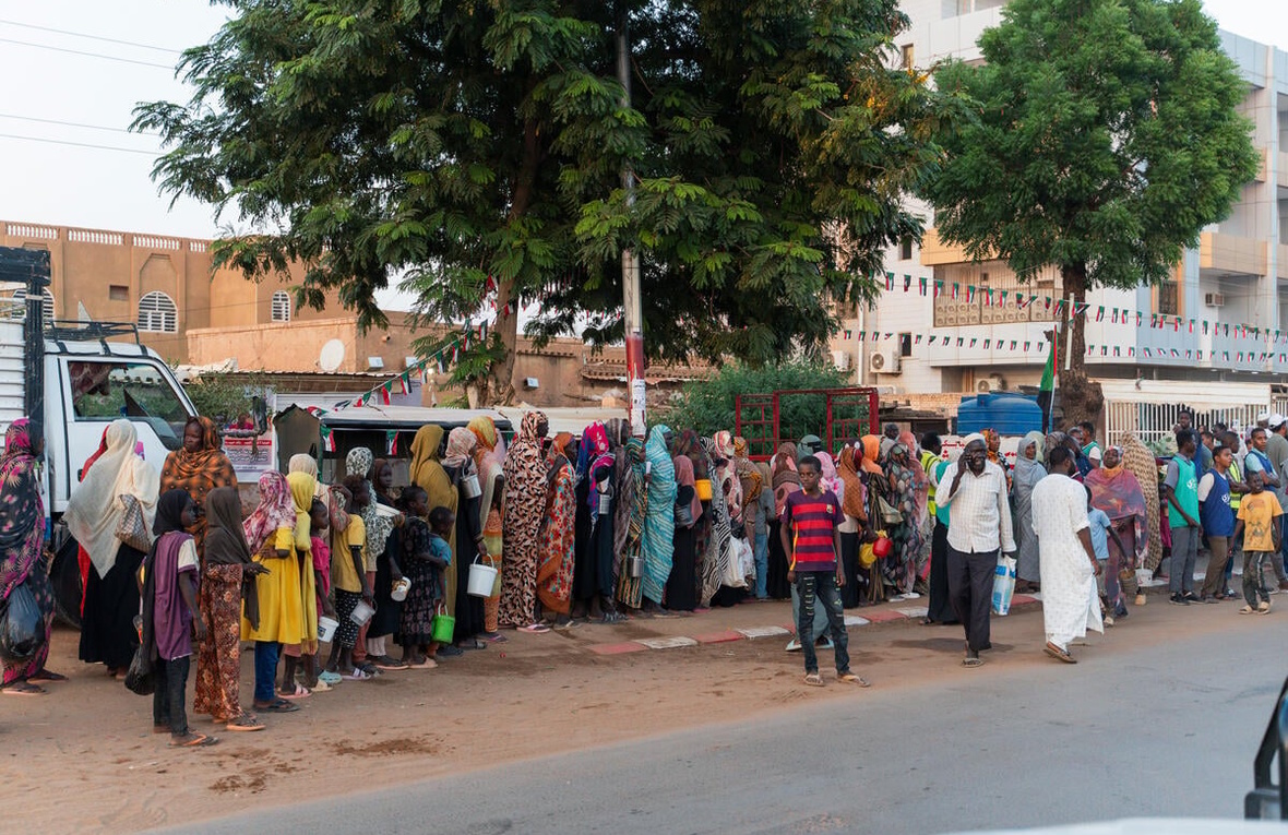 Families from El Fasher queue outside along a street.