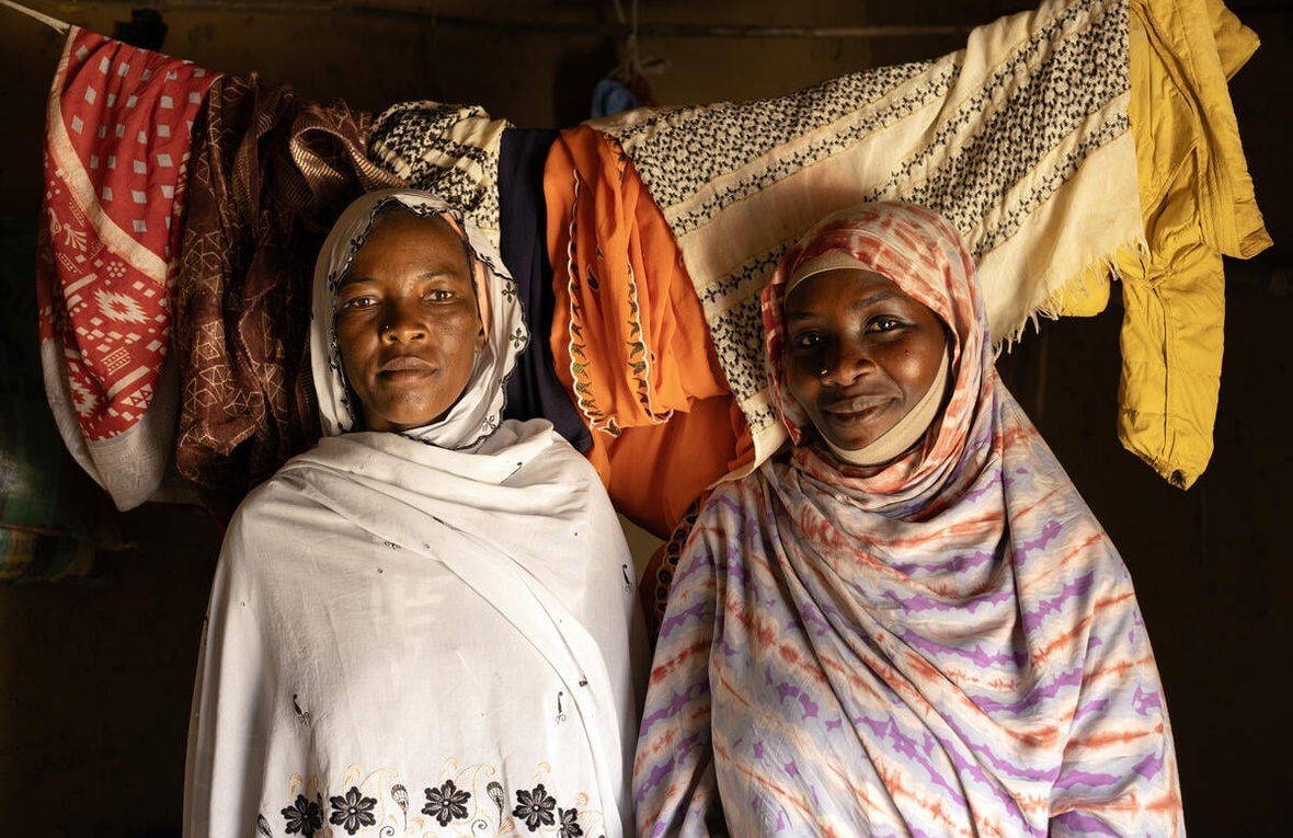 Two women stand next to each other inside a room with washing hanging on a line behind them