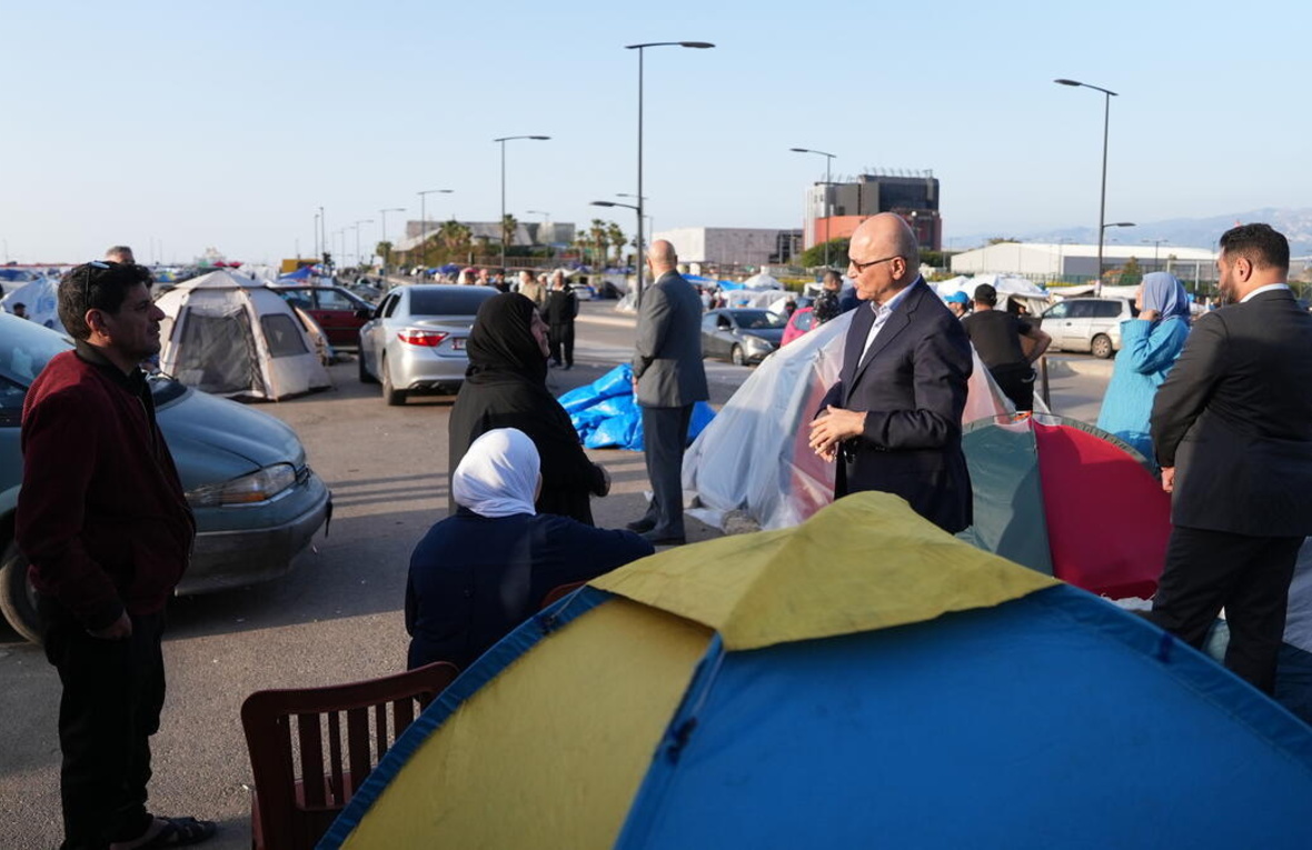 A man in a suit speaks to a family in an open area filled with tents and vehicles 