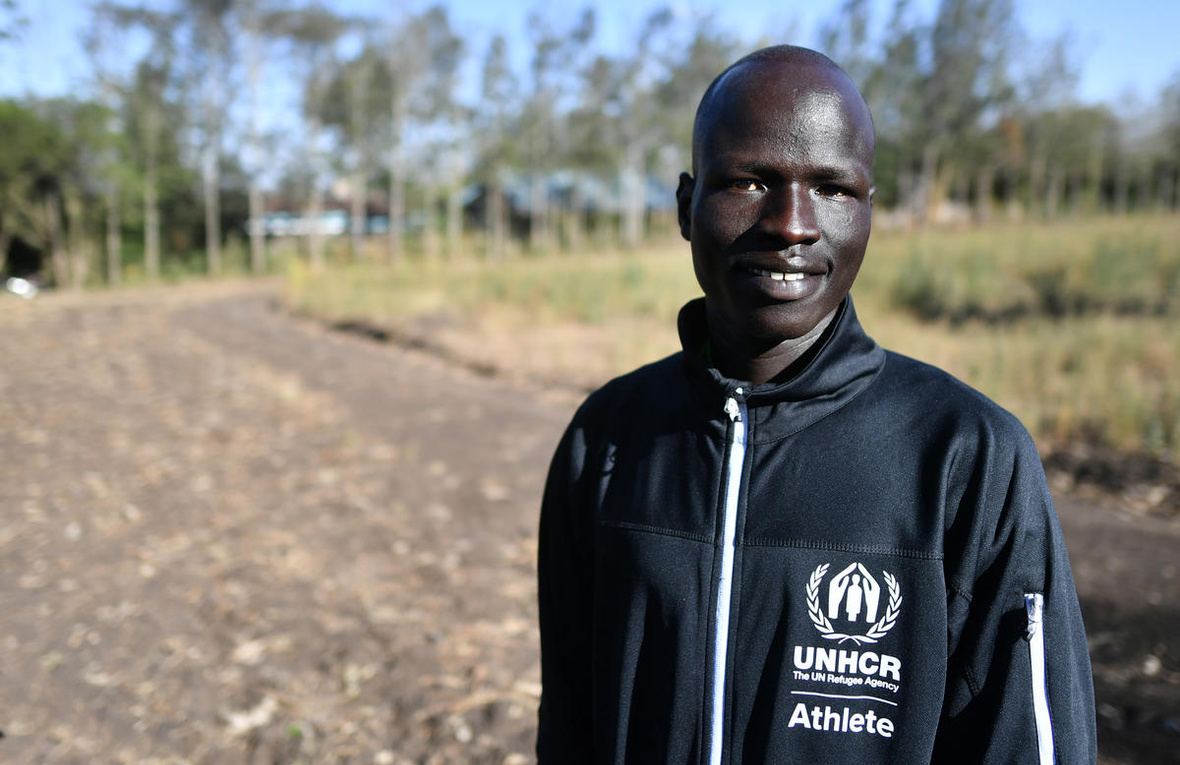 Portrait of a man in a UNHCR-branded tracksuit top standing on a dirt road.
