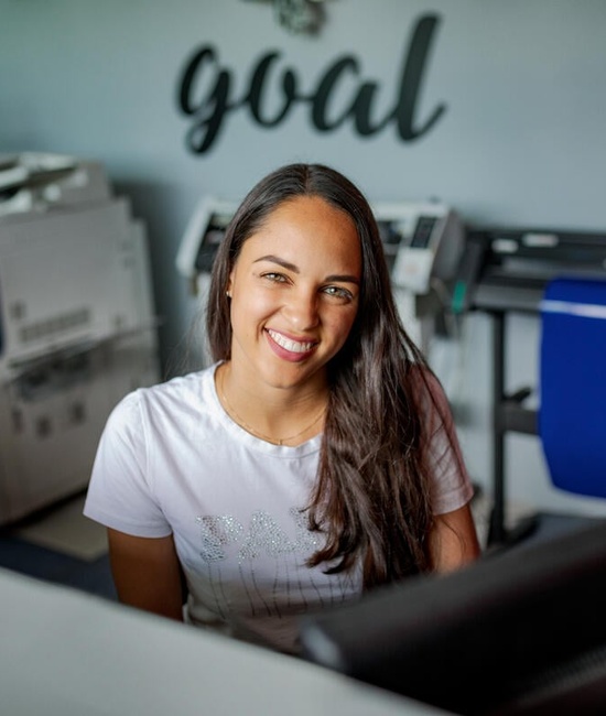 A smiling woman sits behind a desk with a computer in a print shop.