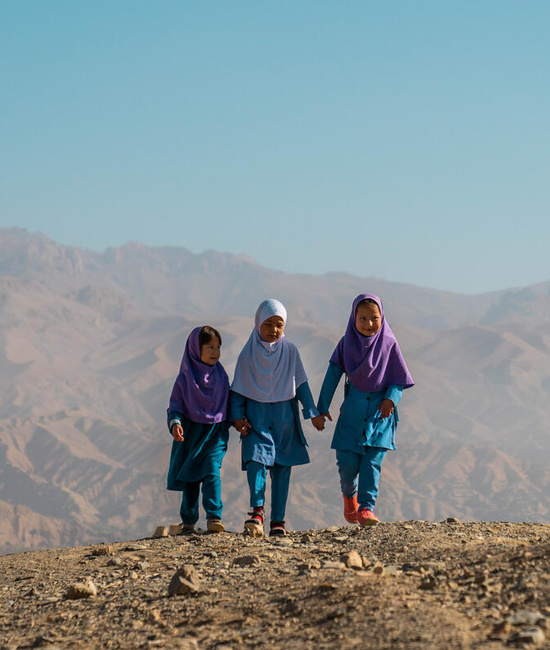 Three young female Afghan refugees walk hand in hand on arid ground, a mountain range behind them