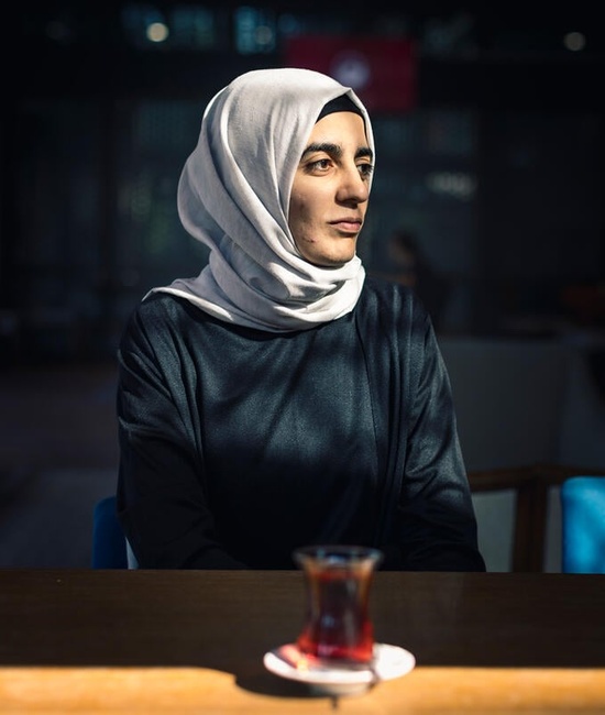 Aicha is surrounded by shadows with her face lit up by the sun as she looks off to the side, a cup of tea on a table in front of her