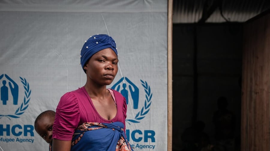 A woman carrying her baby on her back stands in front of a UNHCR shelter.