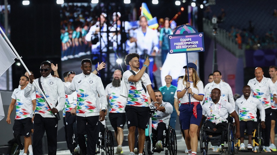 A group of athletes walking and in wheelchairs parade in front of a giant screen display..