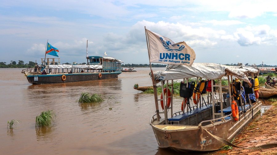 Wooden boats float on a wide, muddy river. One bears a UNHCR flag.
