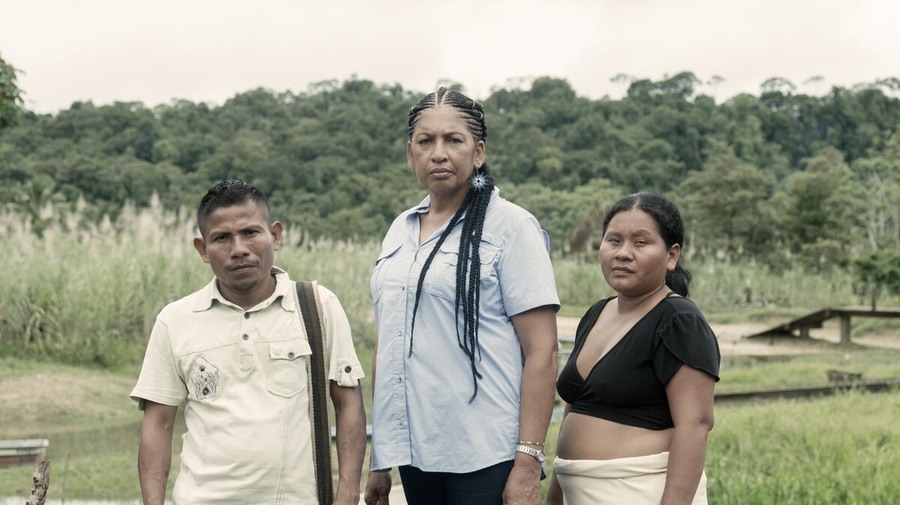 A man and two women stand in front of a stream on the edge of a rainforest.