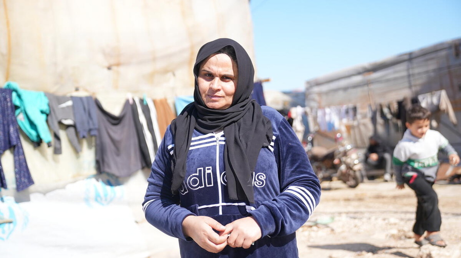 A woman stands outside in front of lines of washing and a UNHCR shelter