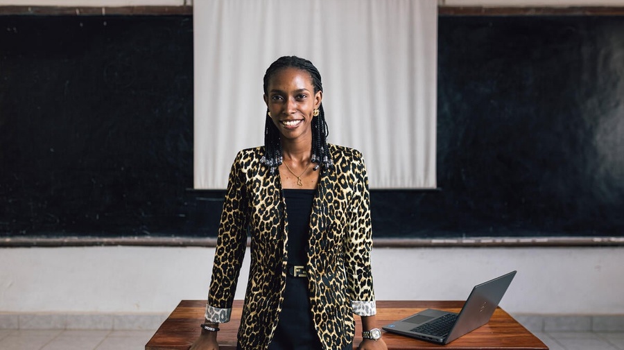 A smiling woman leans against a desk, with a laptop beside her and a blackboard and projector screen behind her