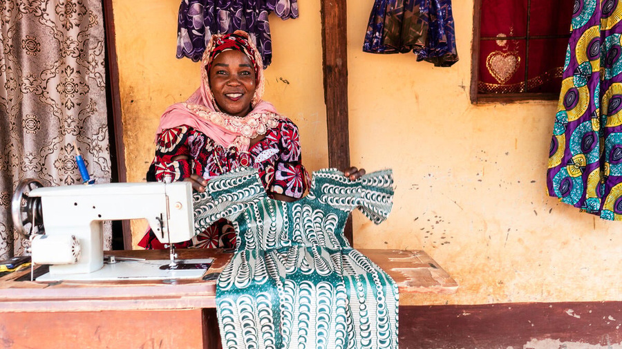 A smiling woman sitting next to a sewing machine table holds up a green and white patterned dress
