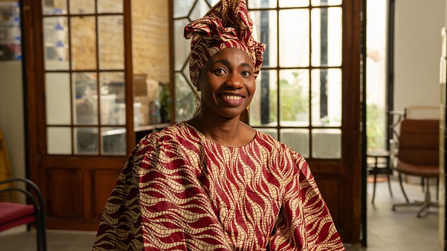 A smiling woman in a patterned traditional dress and headscarft sits in an office