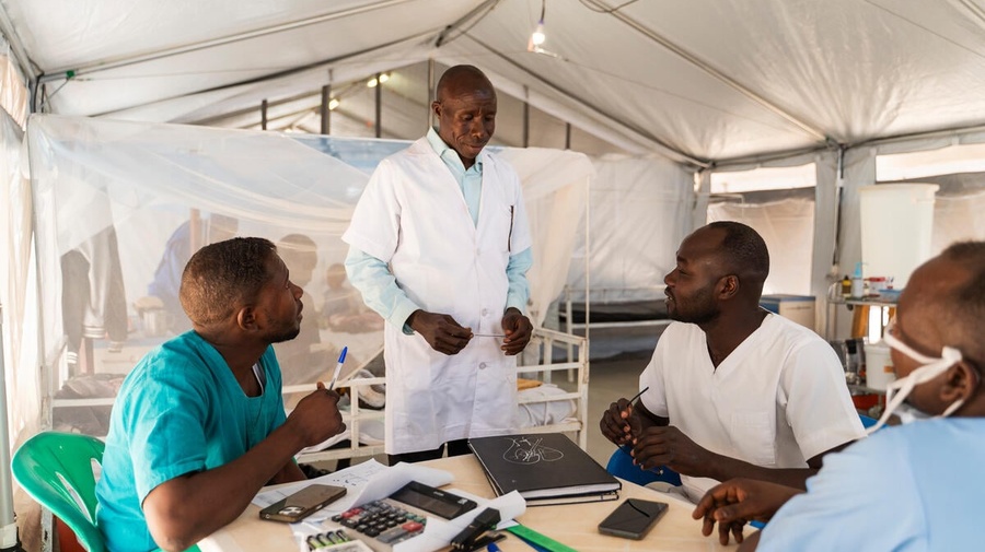 A male nurse in a white coat talks to his colleagues who sit at a round table in a hospital