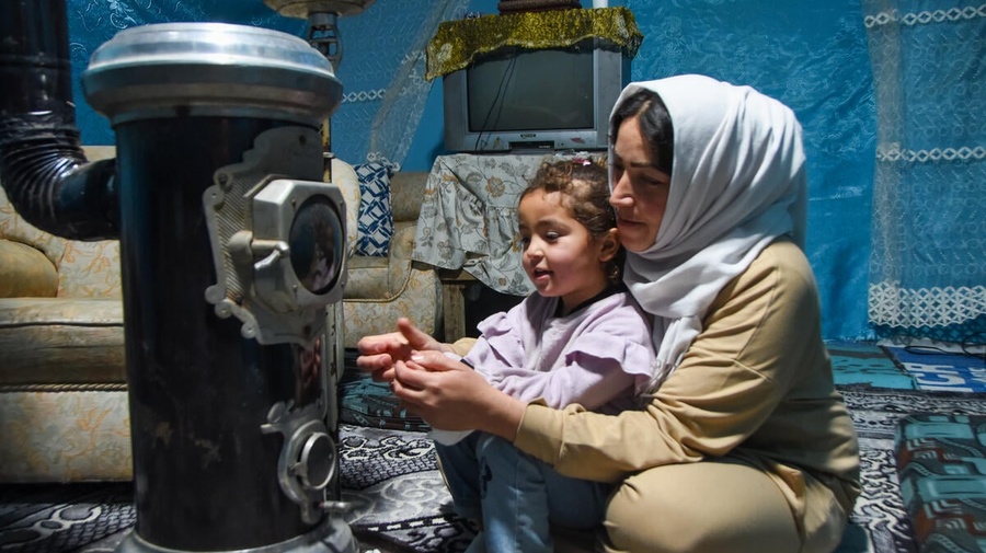 A woman and a young girl warm their hands near an upright stove inside a tented living room