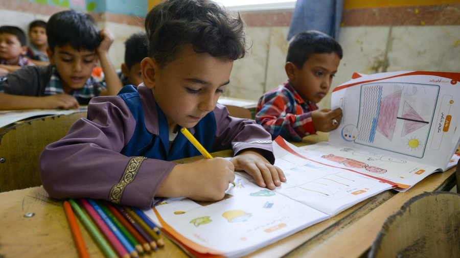 Un niño escribiendo en un cuaderno; a su alrededor, otros niños en un aula