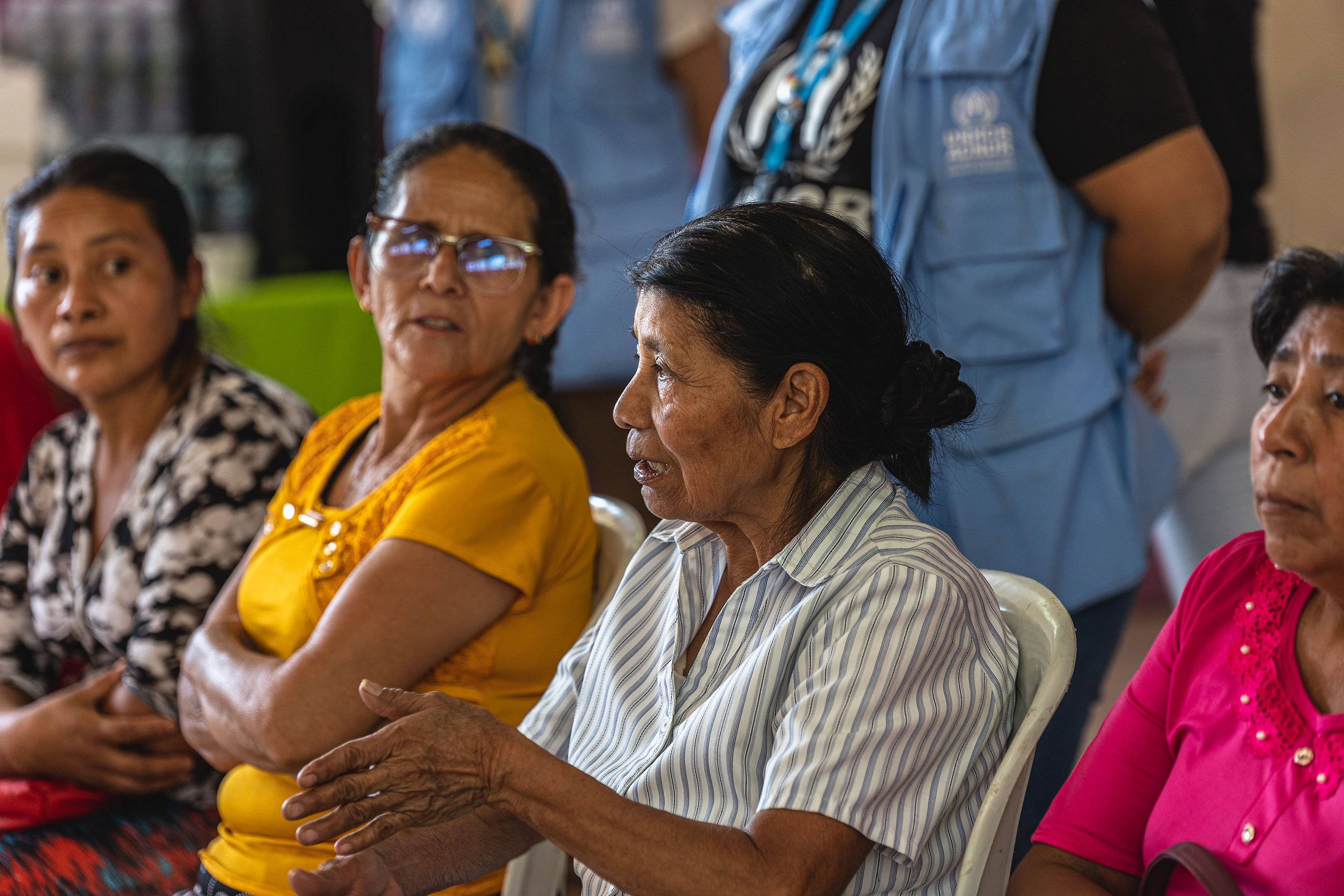 Several older people seated, in a discussion, with UNHCR staff behind them