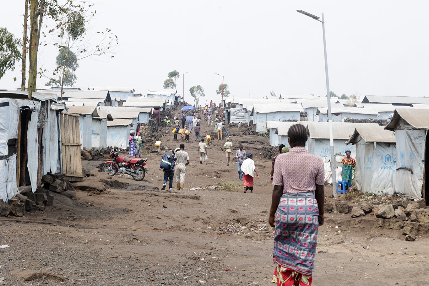 People in ones and twos walk over the undulating dry ground through a camp for internally displaced people