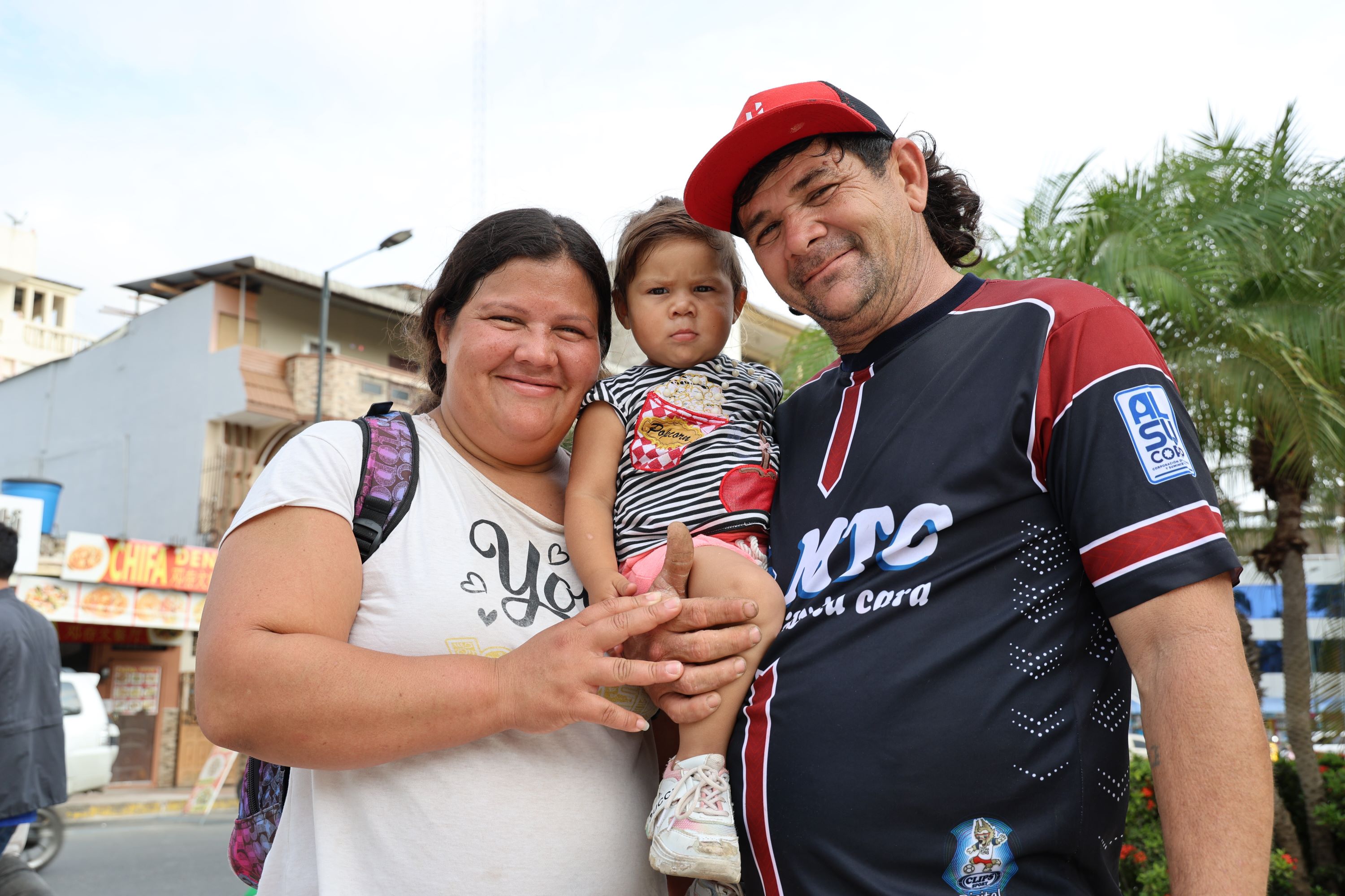 A man and a woman smile at the camera while holding their small child, buildings and a tree in the background.